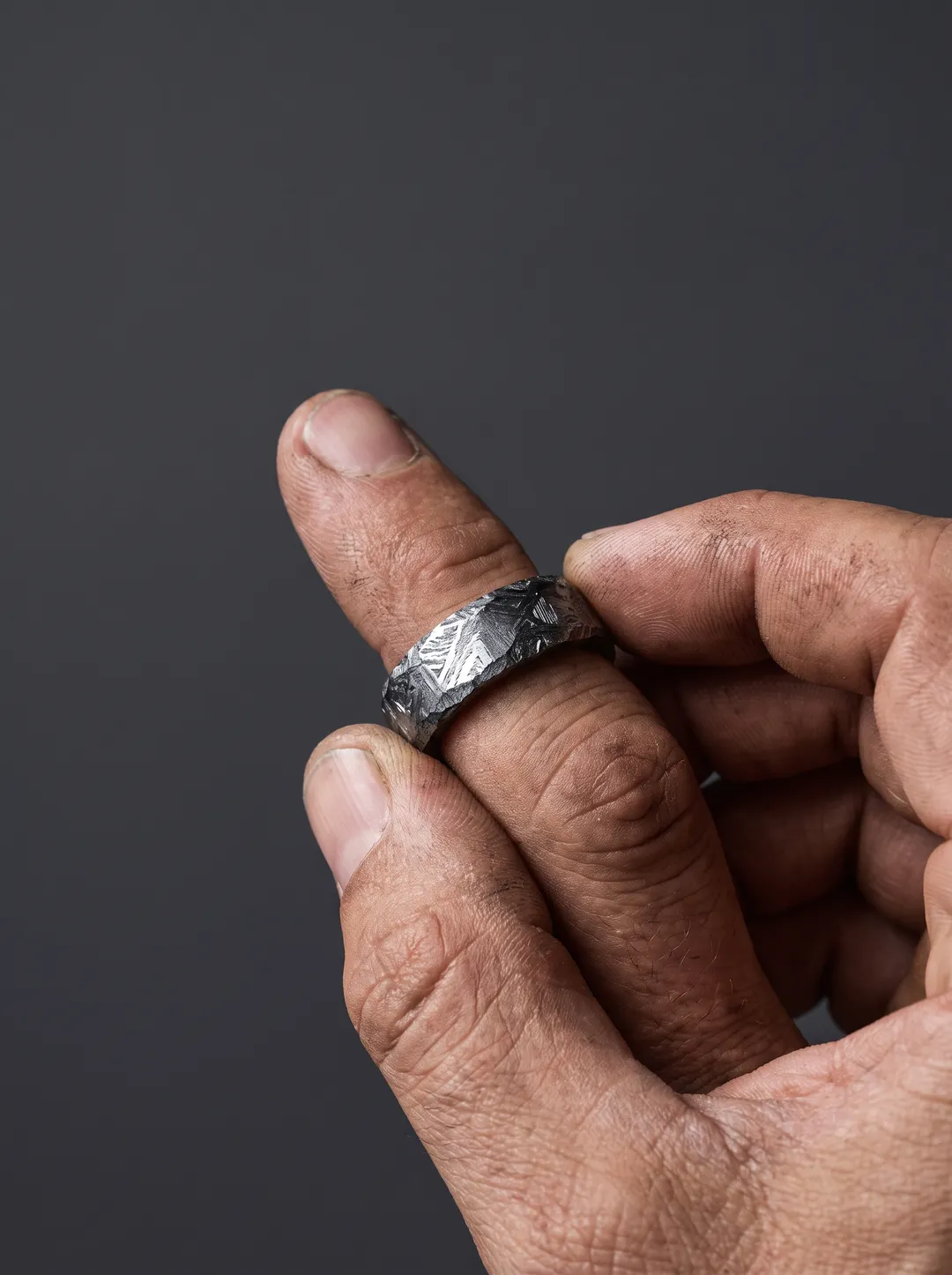Close crop of a weathered hand wearing a wide meteorite iron ring against a solid graphite backdrop