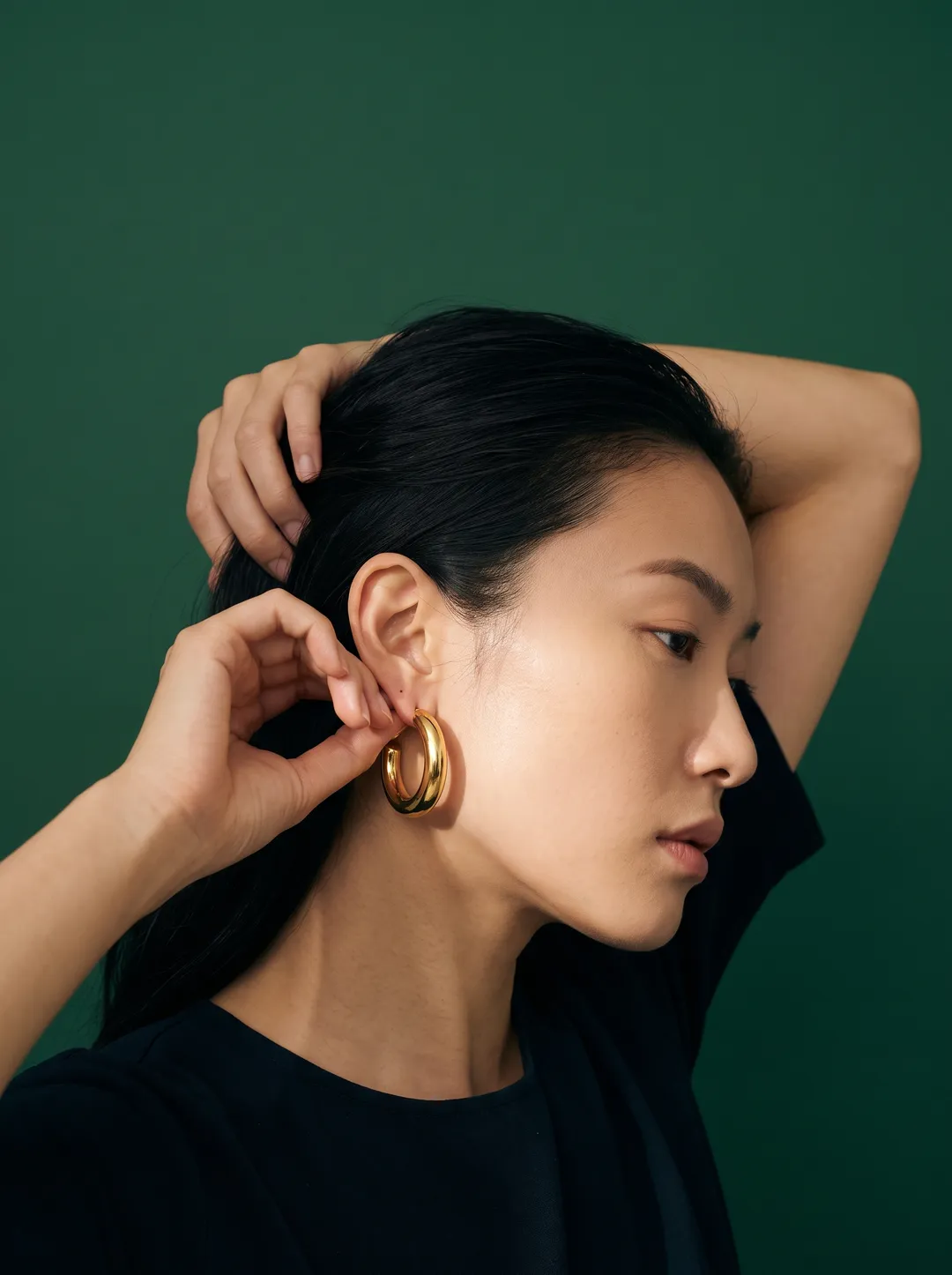 An East Asian woman in profile fastening a gold hoop earring against a solid deep green backdrop