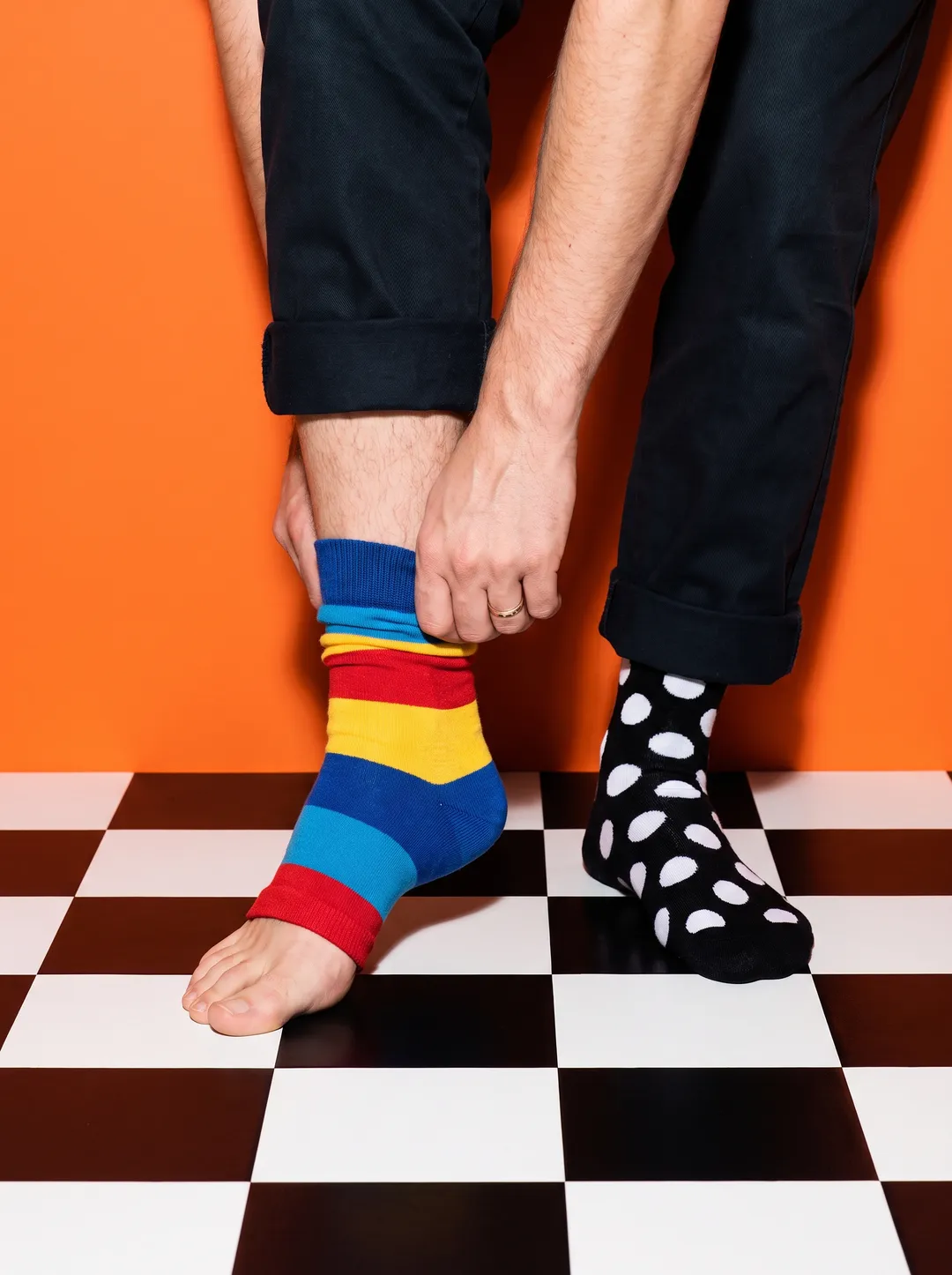 A person's feet on a checkerboard floor pulling on a striped sock against a solid tangerine backdrop