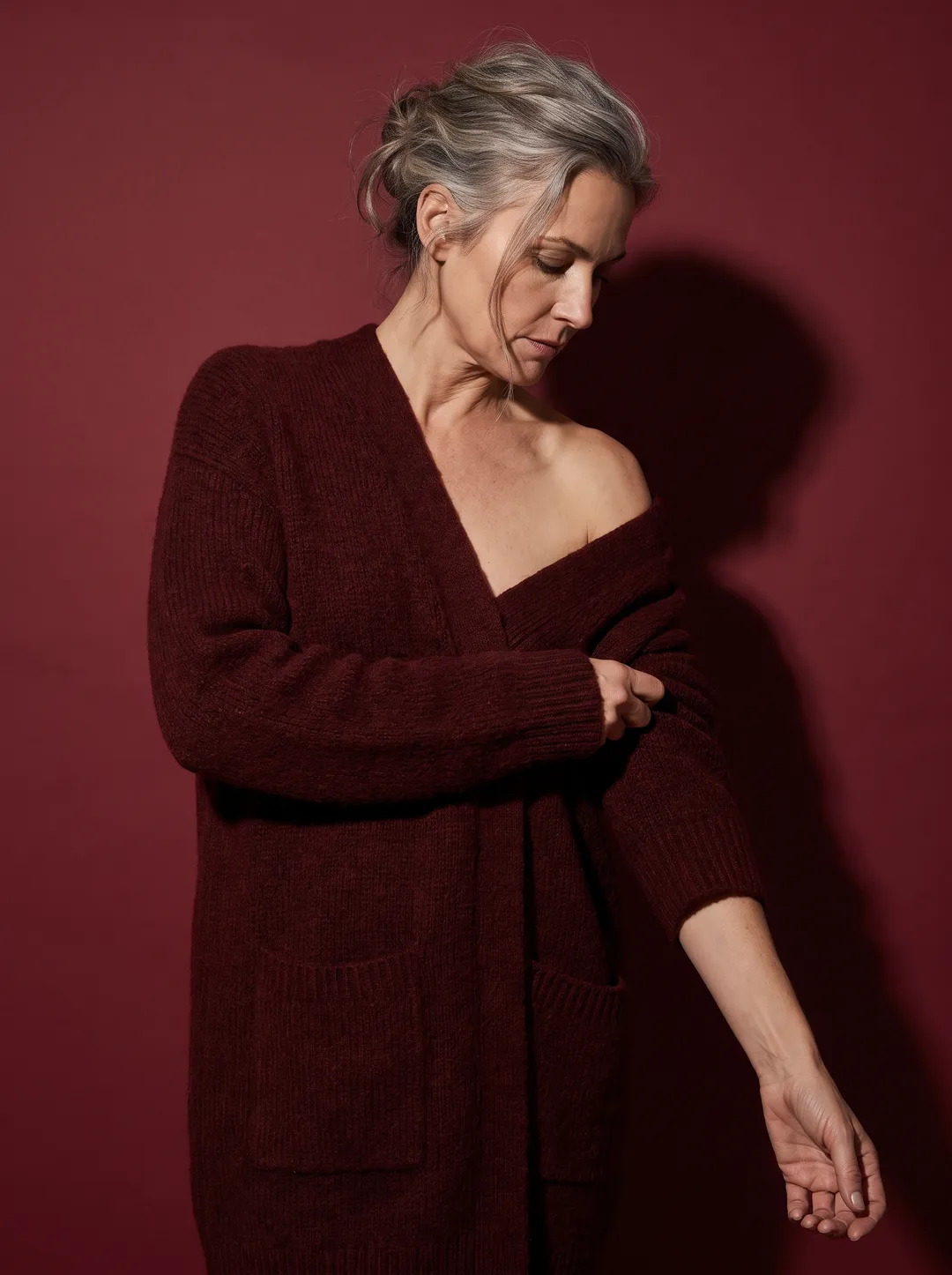 A woman with silver-streaked hair pulling on an oxblood wool cardigan against a solid oxblood backdrop