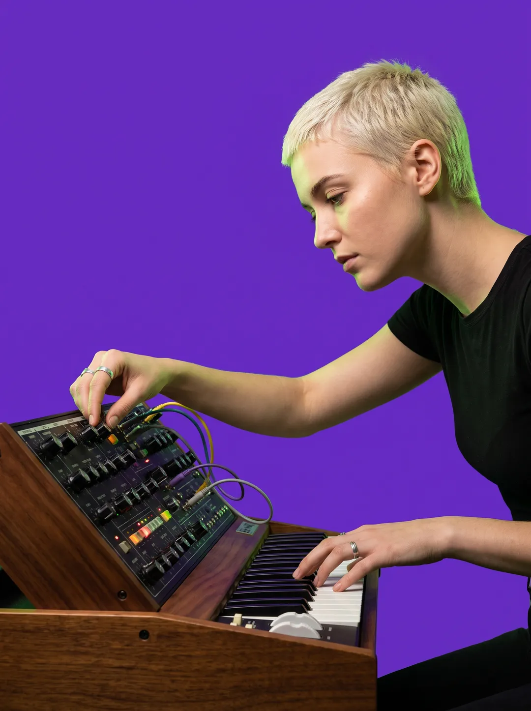 A woman with bleached hair playing a vintage analogue synthesizer against a solid violet backdrop