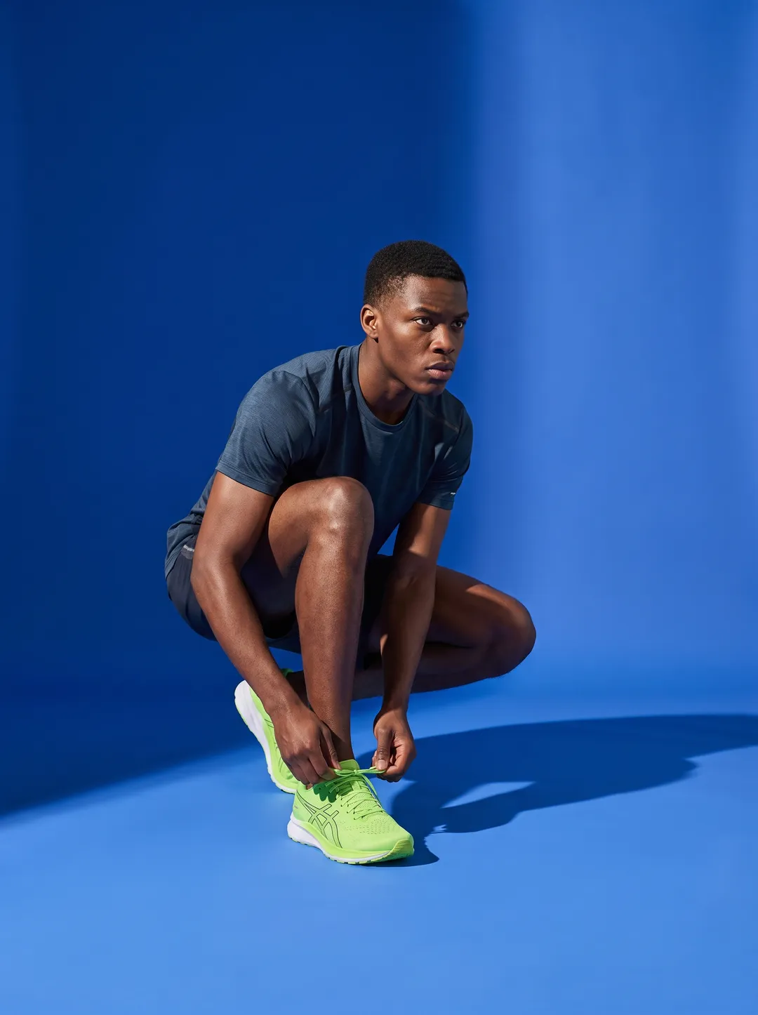 A young Black man crouched tying a lime running shoe against a solid cobalt backdrop