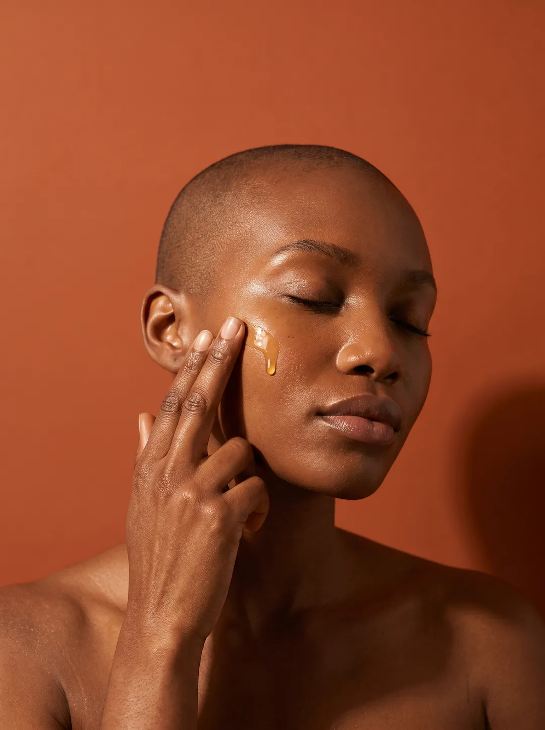 A dark-skinned woman with a shaved head pressing golden serum into her cheek against a solid terracotta backdrop